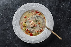 Fresh creamy salmon fish soup with potato, carrots, tomato, peppers and onions in a white ceramic plate on a black background,