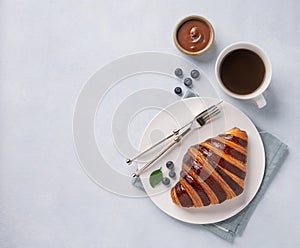 Fresh croissant with chocolate and blueberries on a white plate on a light background with a cup of black coffee. Delicious