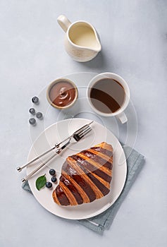 Fresh croissant with chocolate and blueberries on a white plate on a light background with a cup of black coffee and milk jug.