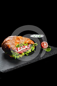 Fresh croissant sandwich with meat and vegetables on black background