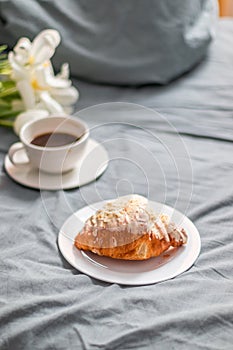 Fresh croissant on a white glass plate and a cup of coffee on the bed. Bouquet of white tulips. Breakfast in bed