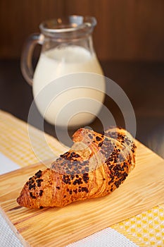 Fresh croissant on a wooden board and kitchen towel with a decanter of milk. Freshly baked croissant on a dark wooden table.