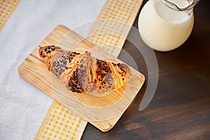 Fresh croissant on a wooden board and kitchen towel with a decanter of milk. Freshly baked croissant on a dark wooden table.