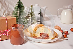 Fresh croissants bun with chocolate on the plate, cup of coffee, jar of milk nearby, three little Christmas tree toy and gift box