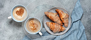 Fresh croissants and hot cups of coffee with hearts on rustic gray background. Top view, flat lay. Wide composition.