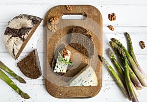 Dark sliced bread, blue cheese with nuts on wooden cutting board decorated with asparagus. Flat lay, top view