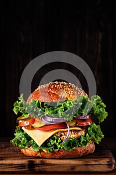 Fresh delicious homemade grilled burger with turkey cutlet with cheese and vegetables on a wooden board, on a dark background.