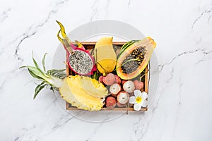 Fresh exotic fruits in wooden box on white marble background - sliced papaya, mango, pineapple, dragon fruit, lychee. Mockup, flat