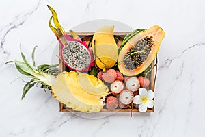Fresh exotic fruits in wooden box on white marble background - sliced papaya, mango, pineapple, dragon fruit, lychee. Mockup, flat