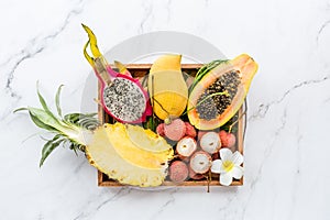 Fresh exotic fruits in wooden box on white marble background - sliced papaya, mango, pineapple, dragon fruit, lychee. Mockup, flat
