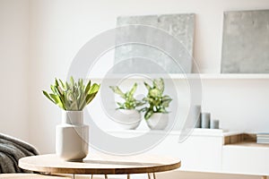 Fresh flowers in white vase placed on small table in bright room interior with paintings, potted plants and candles on shelves in