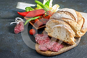 Fresh fuet and sliced homemade bread on the wooden cutting board, fresh vegetables on the dark background