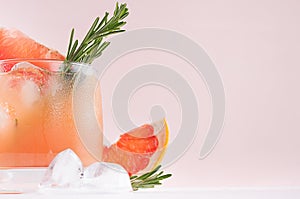 Fresh grapefruit juice with ice, piece and green rosemary closeup on soft white wood table and pink background.