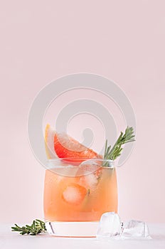 Fresh grapefruit juice with ice, piece and green rosemary closeup on soft white wood table and pink background, vertical.