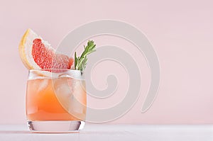 Fresh grapefruit juice with ice, piece and green rosemary on soft white wood table and pink background.
