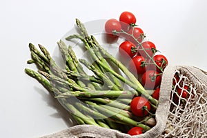 Fresh green asparagus in cotton mesh bag. Fresh cherry tomatoes and green raw asparagus on white background with copy space.