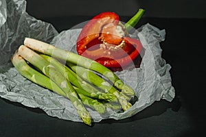 fresh green asparagus and half of red sweet, large pepper lie on white parchment on a dark gray background. proper