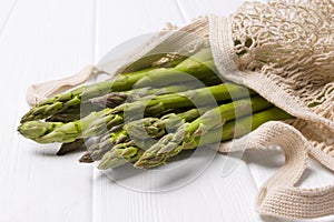 Fresh green asparagus in string shopping bag on white wooden table background
