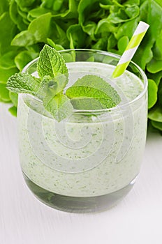 Fresh green spinach smoothie drink with straw, twig mint, salad on soft white wood table, closeup.