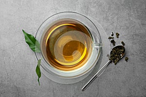 Fresh green tea in glass cup and spoon with dried leaves on grey table, flat lay