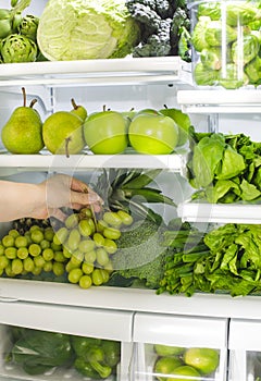 Fresh green vegetables and fruits in fridge. Woman takes the bunch of green grapes from the open refrigerator.