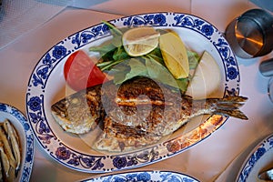 Fresh grilled whole sea bream fish with roasted potato, green salad, tomato and yellow lime on white oval plate with blue pattern