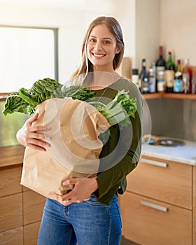 Fresh from the grocers. Portrait of a smiling young woman standing in her kitchen carrying a paper bag full of groceries