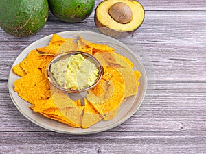 Fresh guacamole with corn tortilla chips in a bowl and avocado halves with seed and whole fruits on a wooden table. Mexican