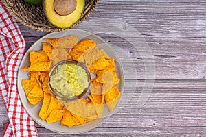 Fresh guacamole with corn tortilla chips in a bowl and avocado halves with seed and whole fruits on a wooden table. Mexican