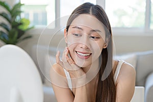 Fresh healthy skin, beautiful smile of asian young woman, girl looking at mirror, applying moisturizer on her face, putting cream