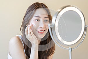Fresh healthy skin, beautiful smile of asian young woman, girl looking at mirror, applying moisturizer on her face, putting cream
