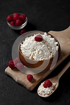 Fresh homemade cottage cheese in a wooden bowl with raspberries on a dark background, healthy food on a black table