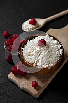 Fresh homemade cottage cheese in a wooden bowl with raspberries on a dark background, healthy food on a black table