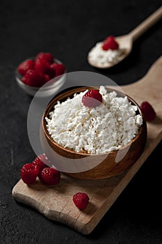 Fresh homemade cottage cheese in a wooden bowl with raspberries on a dark background, healthy food on a black table