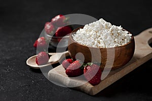 Fresh homemade cottage cheese in a wooden plate with strawberries on a cutting board, healthy breakfast on a black table