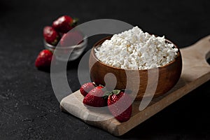 Fresh homemade cottage cheese in a wooden plate with strawberries on a cutting board, healthy breakfast on a black table