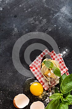 Fresh homemade pasta tagliatelle on wooden spoon with pasta ingredients tomatoes, raw egg, basil leaf on the dark concrete backgro