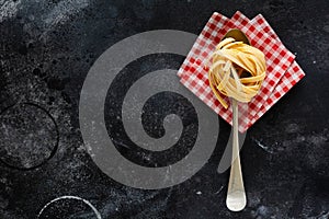 Fresh homemade pasta tagliatelle on wooden spoon with pasta ingredients tomatoes, raw egg, basil leaf on the dark concrete backgro