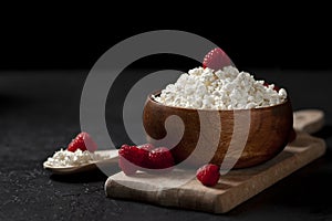 Fresh homemade pure cottage cheese in a wooden plate with raspberries in a rustic bowl against a dark background, healthy food on