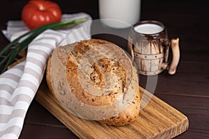 Fresh homemade rye bread, tomato, bunch onion, clay mug and bottle of milk on wooden cutting board