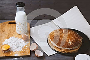 Fresh, hot pancakes in a frying pan, eggs, milk, flour on a wooden table.