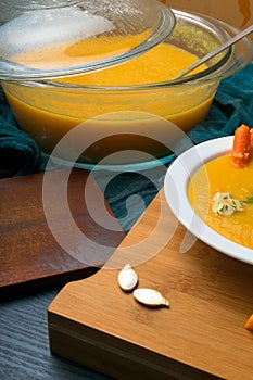 fresh hot pumpkin soup in glass pan and in white plate, decorated with mini carrot and pumpkin seeds
