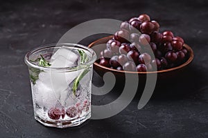 Fresh ice cold carbonated water in glass with rosemary leaf near to wooden bowl with grape berries, dark stone background