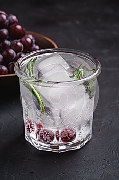 Fresh ice cold carbonated water in glass with rosemary leaf near to wooden bowl with grape berries, dark stone background