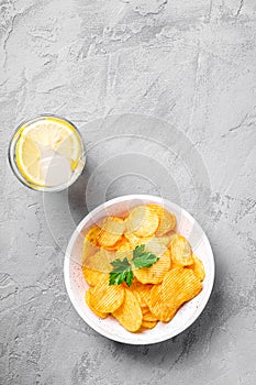 Fresh ice cold water drink with lemon near to golden potato chips with parsley leaf in wooden bowl on concrete background