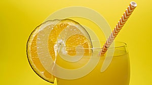 Fresh juice with a slice of orange on a glass and a straw, a summer drink on a bright yellow background, a close-up of orange