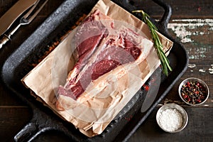 Fresh, juicy seasoned t-bone steak on food paper in a grill pan with sprinkled spices, rosemary and salt and pepper peas nearby.