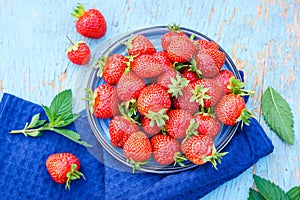 fresh juicy strawberries in a blue plate on an old wooden rural table, next to it is a dark blue napkin and mint leaves