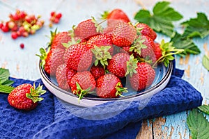 fresh juicy strawberries in a plate on an old wooden rural table, next to it is a dark blue napkin and currant berries