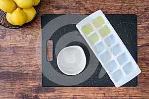 Fresh lemon juice in an ice cube tray, black cutting board, wood table, basket of lemons, white bowl